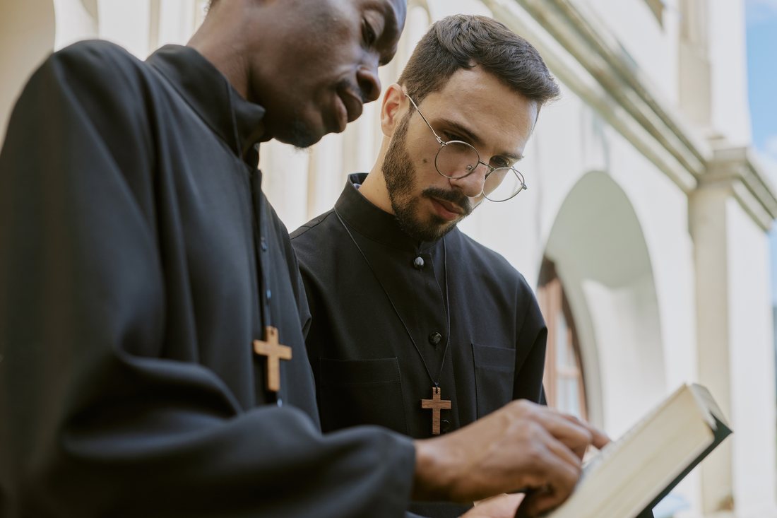 Two men in black cassocks with crosses reading a book together