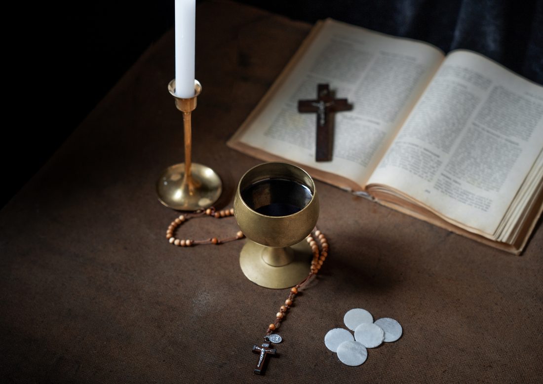 An open Bible, chalice, rosary and communion hosts on a table