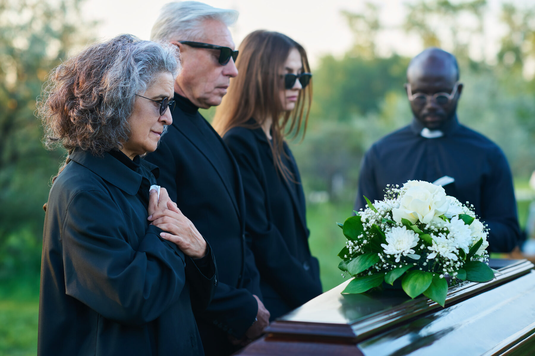 Mourners and a priest beside a casket with white flowers